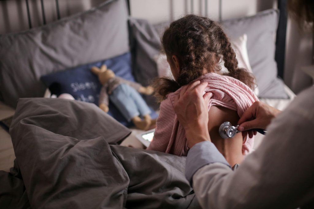 A pediatric doctor checks a child's back with a stethoscope in a cozy bedroom setting.